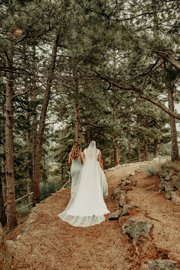bride and bridesmaid walking down the nature aisle surrounded by evergreens to the ceremony site where the groom and guests await at the Sunrise Amphitheater in Boulder on a beautiful summer afternoon.