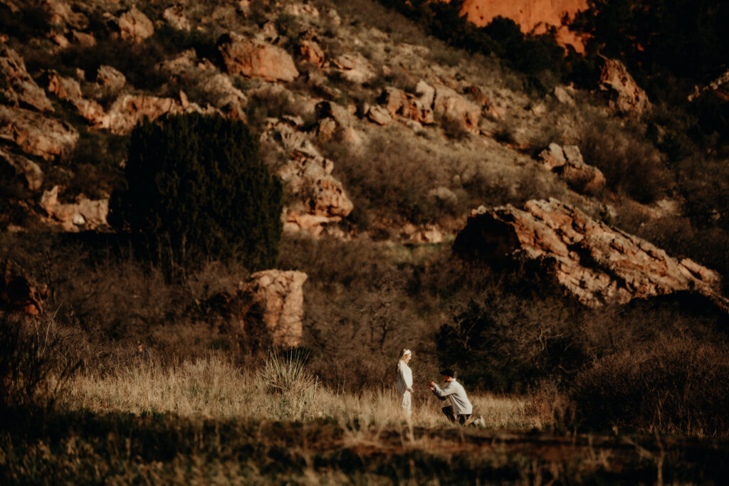 "Nick kneeling to propose to Callie on a quiet trail at Garden of the Gods, captured from a distance with a telephoto lens."