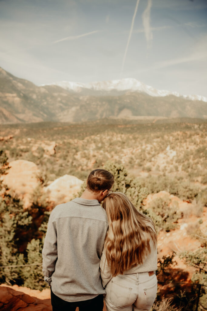 Nick and Callie at the Garden of the Gods looking off into the distance, snuggling with Pikes Peak snowy cap in the background