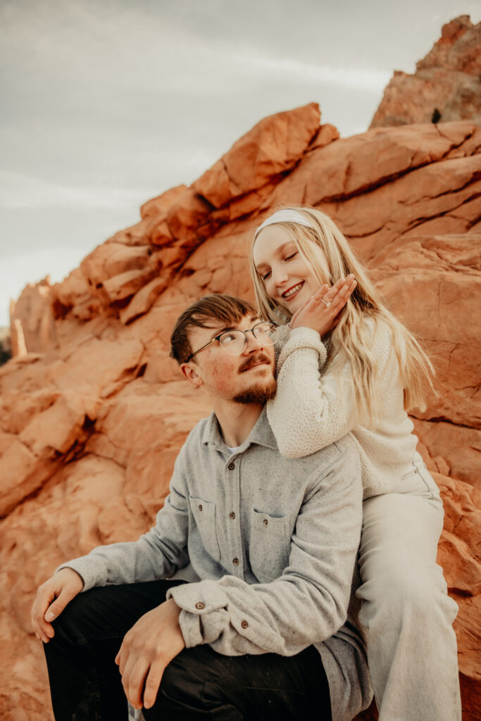 Sitting down pose with Callie resting her elbows on Nicks shoulder showing off her ring as she frames her face and they smile at each other at the Garden of the Gods