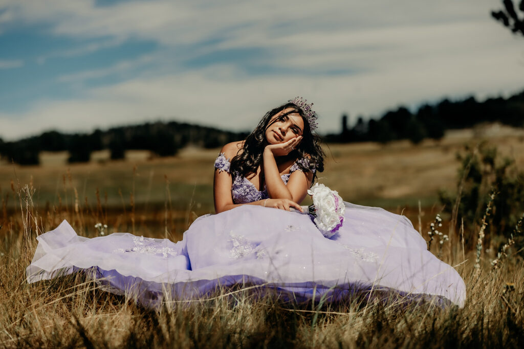 Quinceañera portrait of Natalia in a lavender purple ball gown sitting with a tiara in an open field in Evergreen, Colorado