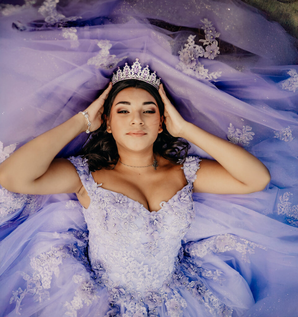 Quinceañera portrait of Natalia in a lavender purple ball gown laying with her hands on the tiara in an open field in Evergreen, Colorado