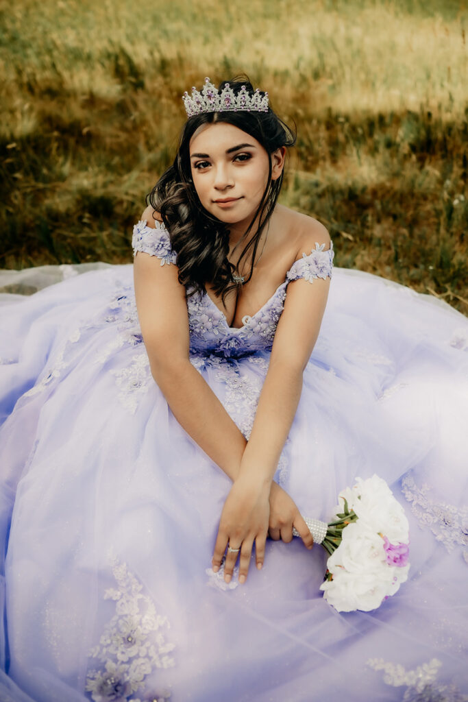 Quinceañera portrait of Natalia in a lavender purple ball gown sitting with a tiara in an open field in Evergreen, Colorado
