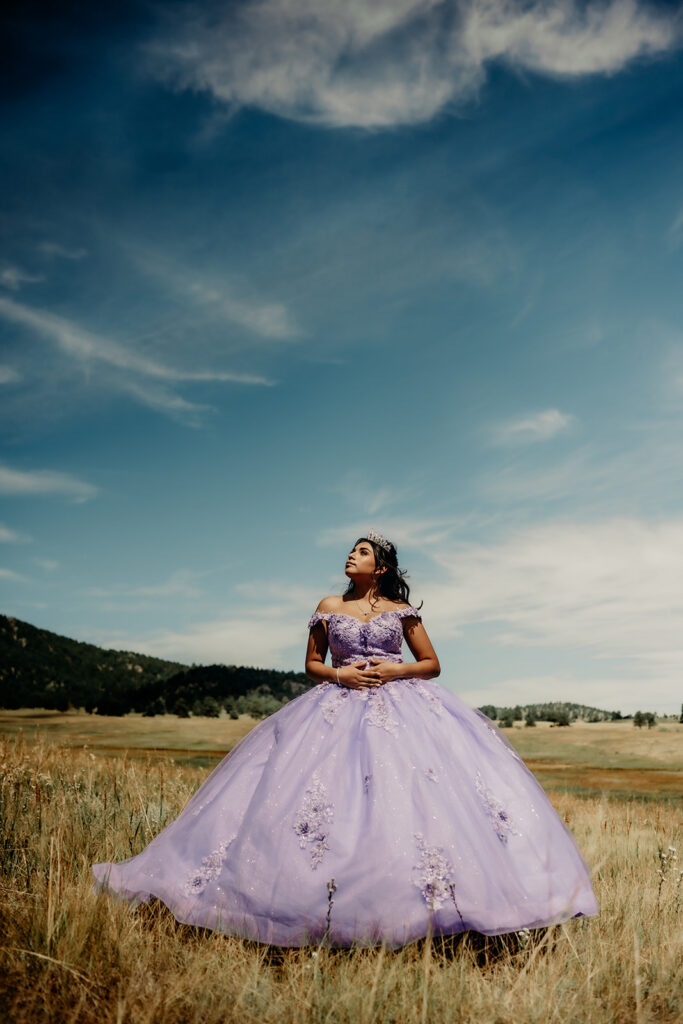 Quinceañera portrait of Natalia in a lavender purple ball standing with her hands resting on her stomach in an open field in Evergreen, Colorado