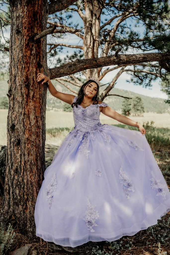 Quinceañera portrait of Natalia in a lavender purple ball standing leaning against a tree in an open field in Evergreen, Colorado