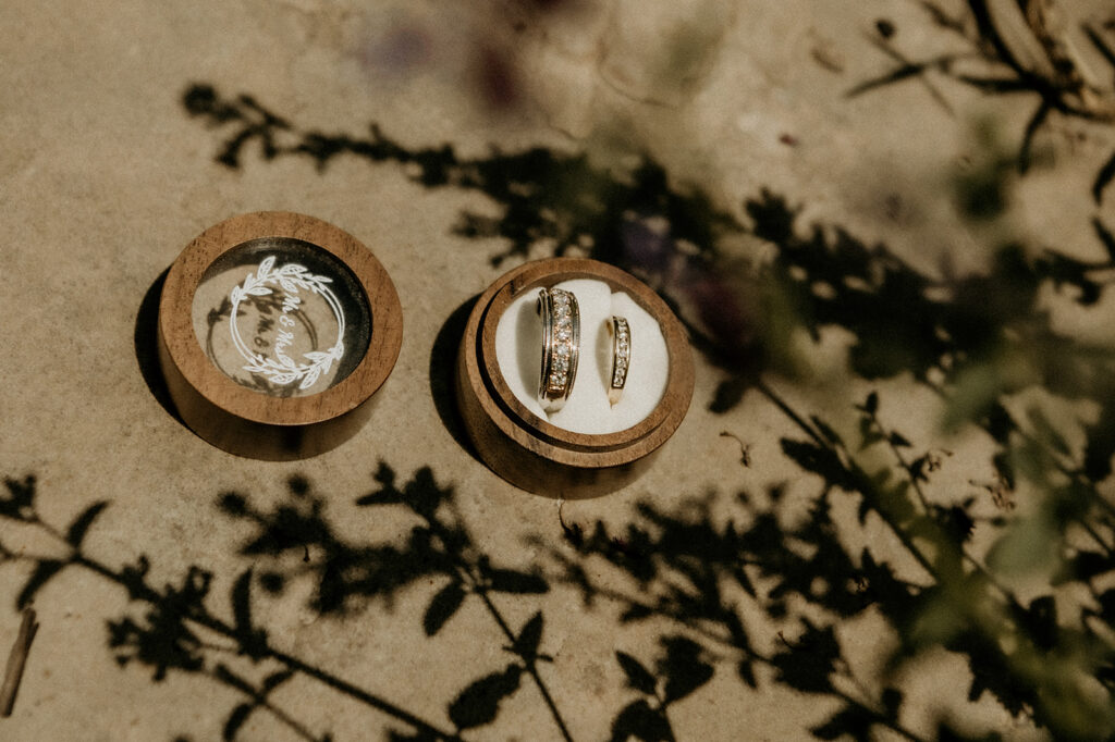 Wood ring box featuring his and her wedding bands full of diamonds featured on the ground framed by plant shadows