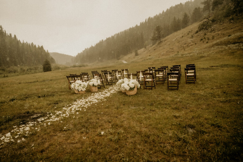 white floral in big tan woven baskets for the ceremony white white petals on the ground creating the aisle way. Ceremony set up in the valley of the mountain ranch in the rain